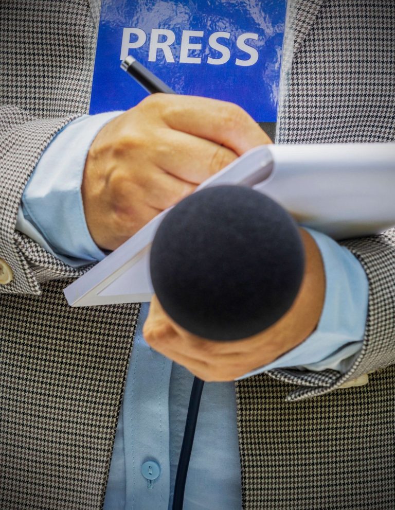 A person holding a microphone and notepad, with a "PRESS" badge visible.