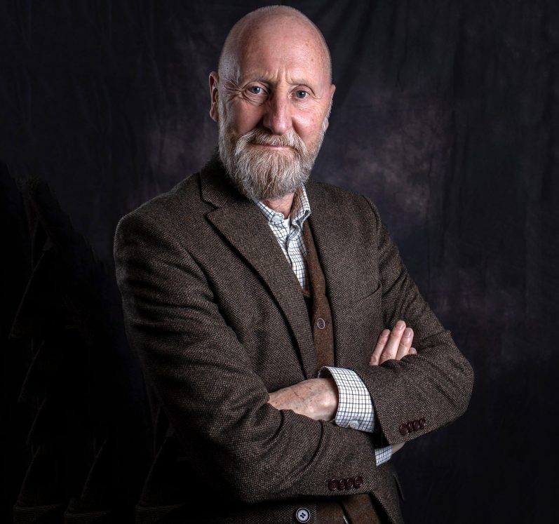 A bearded man in a brown blazer poses with arms crossed, smiling at the camera.