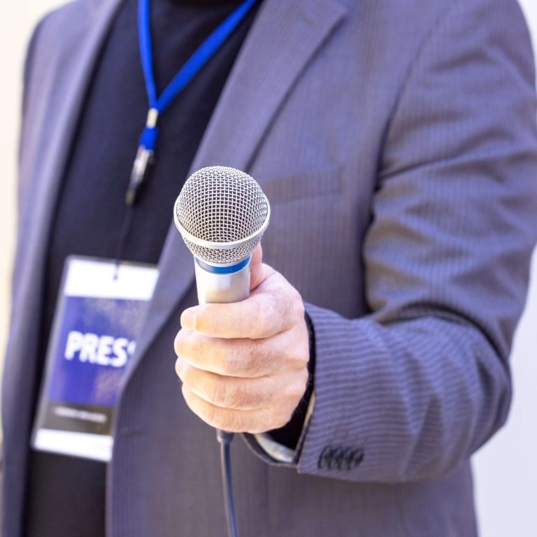 A person holding a microphone, wearing a press badge and a blue lanyard.